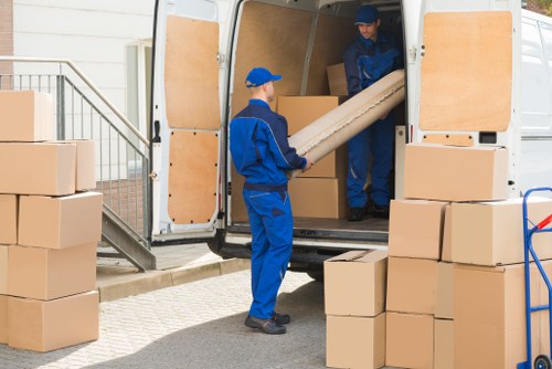 Two removal workers dressed in blue uniforms are loading a cardboard box into the back of a white removals van parked on a residential street near a house with a white exterior and steps. The worker inside the van is passing the box to the worker standing outside, who is holding a long tube-shaped item. Several stacked cardboard boxes of varying sizes are positioned on the ground around the van, some on a hand truck, indicating a house move. The van's interior is visible, with wooden paneling lining the sides. Natural daylight illuminates the scene, suggesting a clear day. The setting is typical of a local house removal in Bow, with a focus on efficient handling of household items including furniture, boxes, and protective materials, consistent with professional removals services offered by Bowmanwithvan.co.uk.