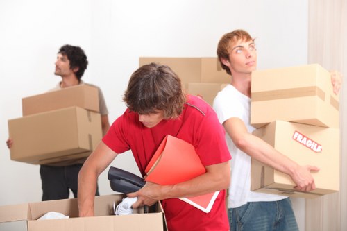 Two men are engaged in house moving activities inside a residential property, with a plain white wall and closed curtains visible in the background, suggesting a room in a UK home. The man in the foreground, wearing a red shirt, is bending over a large open cardboard box while holding a cordless drill and some documents or folders, indicating packing or unpacking tasks. His focus is directed toward the box, which contains various household items. Beside him, another man with light brown hair, dressed in a white t-shirt, is standing upright and carrying a medium-sized cardboard box labeled 'Fragile', with visible packing tape securing it. Behind these two, a third man with dark hair and a grey shirt is visible, holding a larger box in each hand, looking away to the left. The scene captures a typical house removal preparation, with cardboard boxes of different sizes, some stacked and others being transported, emphasizing the process of packing or unpacking during a local move in the Bow area. The environment reflects a residential space used for a professional domestic move, aligned with the services offered by Bow Man with Van.