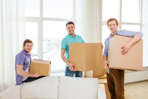The image shows three men with light skin and smiling expressions in a bright, modern room with large windows that allow natural daylight to fill the space. They are engaged in a house removal or relocation activity, each holding cardboard moving boxes of various sizes. The man on the left, dressed in a purple shirt, is seated on a white upholstered sofa, holding a small cardboard box. The man in the middle, wearing a turquoise polo shirt, stands in the center and is lifting a medium-sized box, with a background of large windows revealing a residential area. The man on the right, dressed in a light blue shirt and brown trousers, stands near a radiator and holds a larger cardboard box, appearing to prepare for loading or unloading. The room contains minimal furniture, emphasizing the focus on the movement of household items, with the ceiling and wall reflections indicating a contemporary interior design. This scene depicts a professional house removal service, consistent with the expertise provided by Bowman with Van in the Bow area, promoting local house removals and quotations accessible to residents in postcode area related to Bow, London.