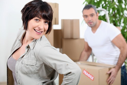 A woman with short dark hair and a light grey jacket smiling at the camera while standing in a room filled with cardboard boxes of various sizes, some stacked on top of each other. Behind her, a man with short dark hair wearing a white t-shirt is also visible, holding a medium-sized cardboard box labeled with a red sticker. The background includes a partially open doorway and a green leafy plant, indicating a residential interior. The environment appears well-lit and tidy, with furniture or household items that suggest the scene is part of a house or flat move. The image reflects a professional house removals service, such as those offered by Bowman With Van, operating within the Bow or nearby area, supporting local moves from a property in the vicinity of Bow, London.