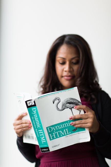 A woman with dark, wavy hair, wearing a black blazer over a burgundy top, is standing indoors holding an open textbook titled 'Dynamic HTML' with a cover illustration of a flamingo. She is focused on 