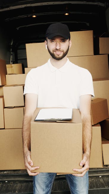 A white moving van parked on a narrow street in a residential area during daylight, with a large, covered sofa and several cardboard boxes secured inside the vehicle, visible through the open rear doo